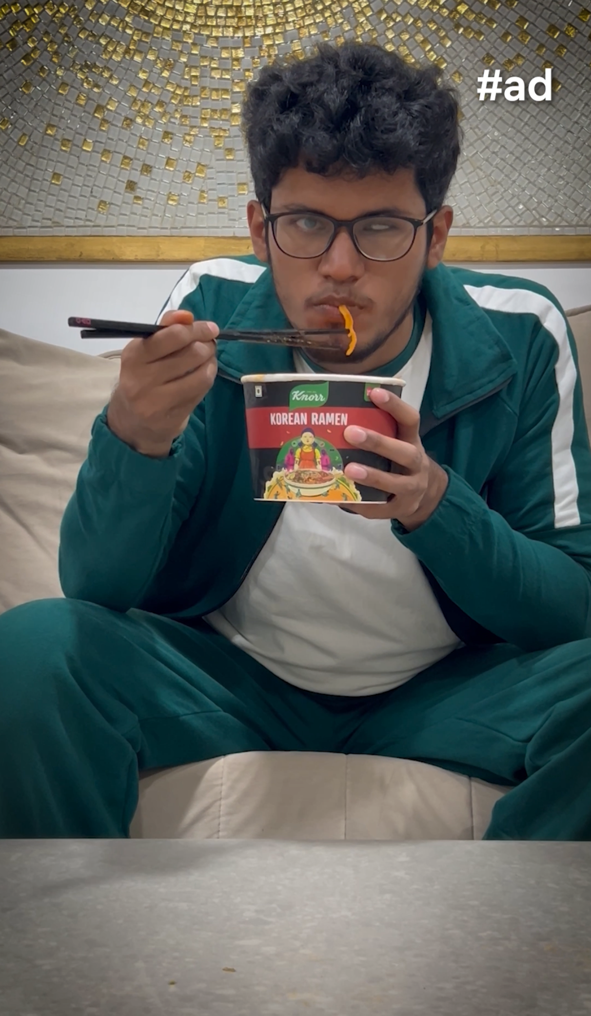 A sponsored ad featuring Bhavya in a green tracksuit, sitting on a couch while eating a cup of Knorr Korean Ramen with chopsticks